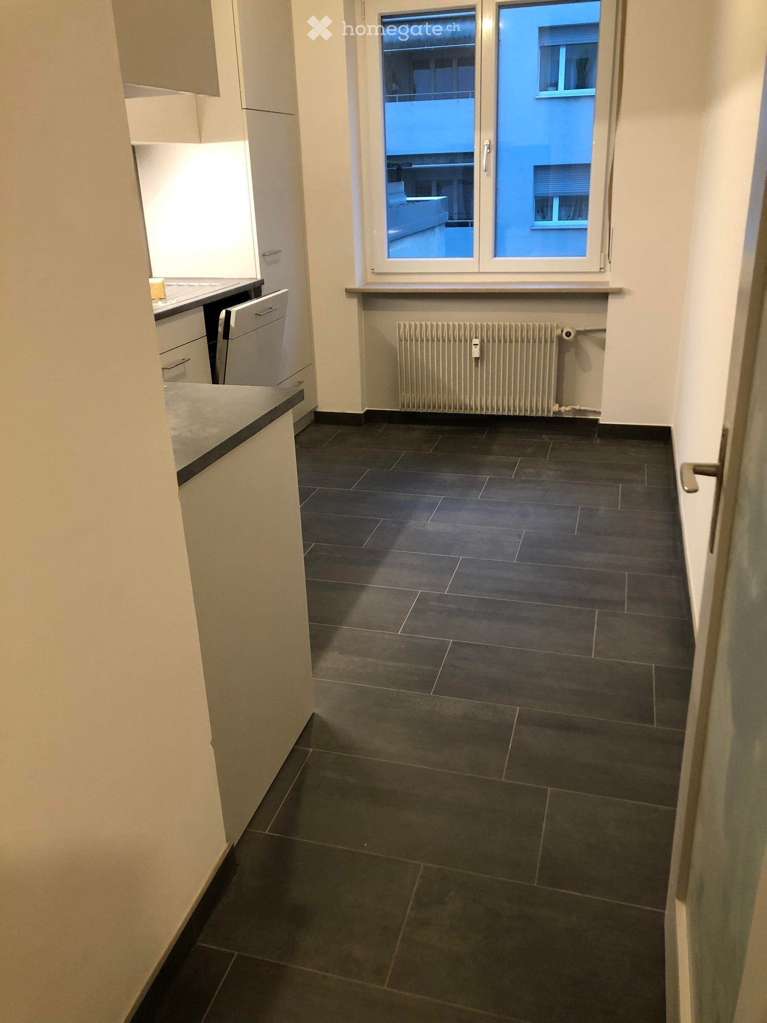 Empty room with dark tiled floor, white walls, radiator, and a window with a view of a balcony area.