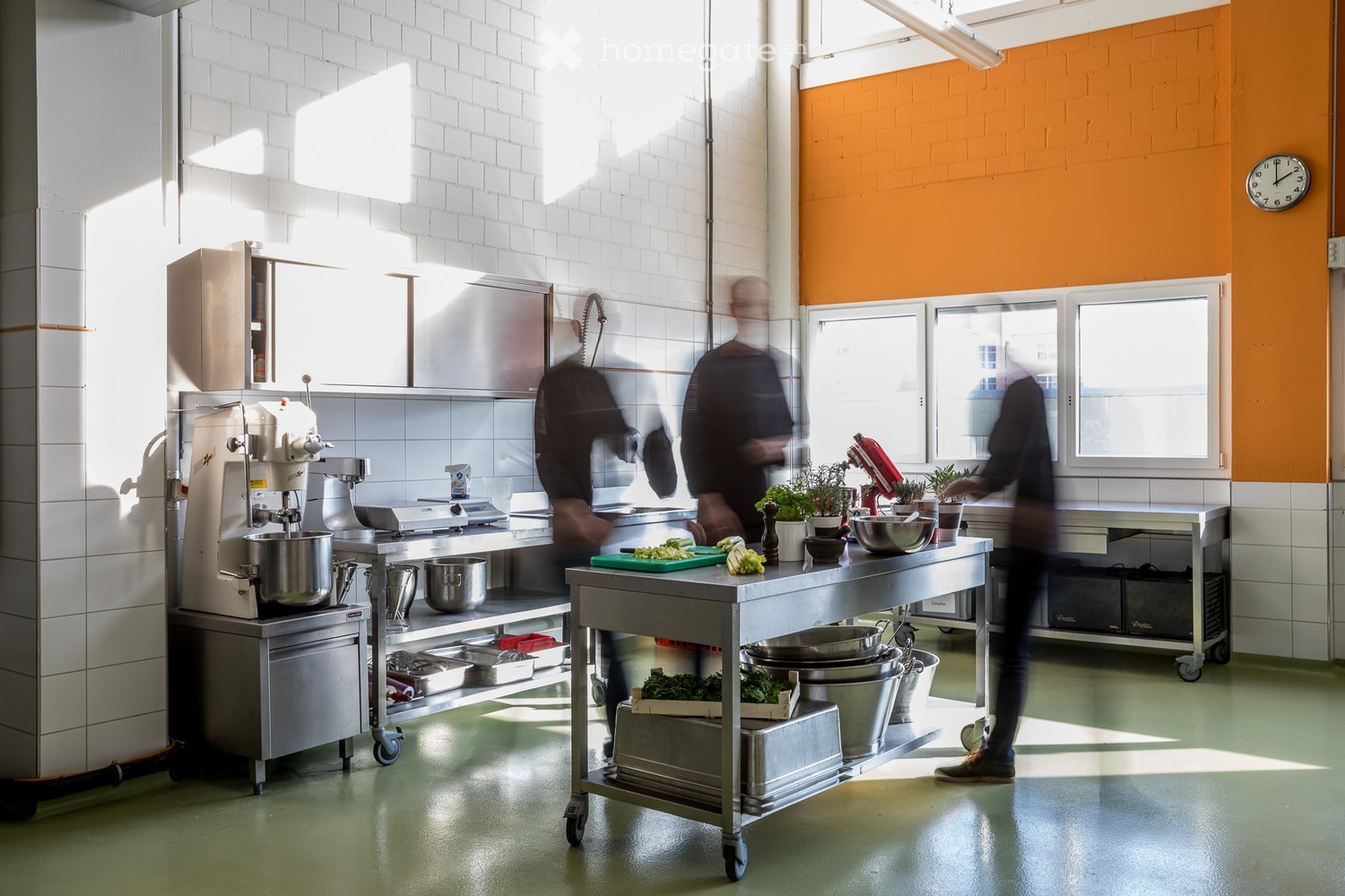 Commercial kitchen with stainless steel appliances, mixer, bowls, cutting board, potted plants, and a clock on the wall.