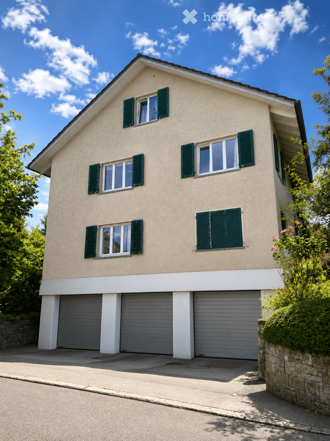 Two-story house with beige siding, green shutters, multiple windows, and a three-car garage.
