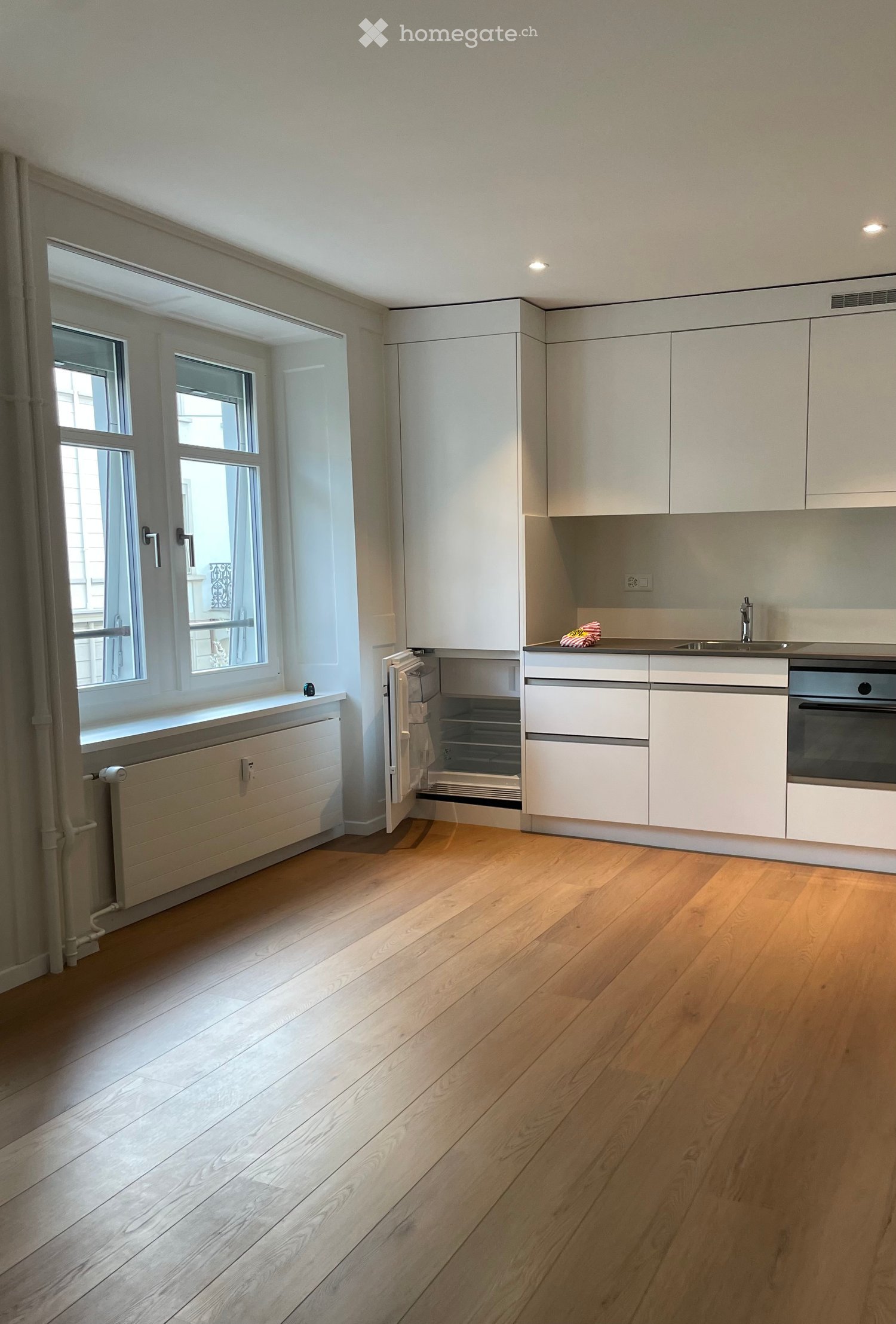 Modern kitchen with light-colored cabinets, wooden flooring, and large windows allowing natural light. Features include a sink, faucet, oven, and refrigerator.