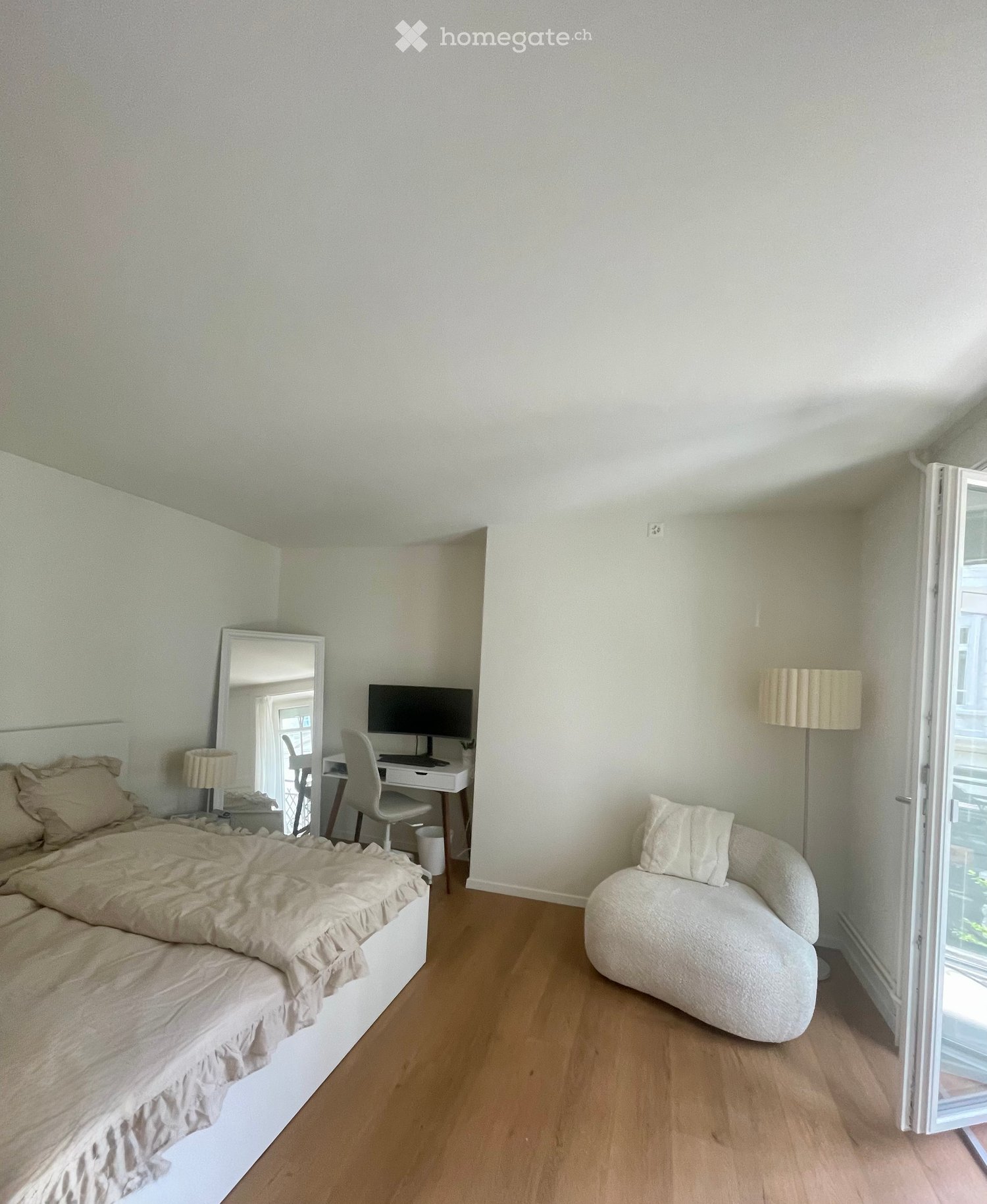 A well-lit bedroom with a bed, a sofa, a desk with a chair, a mirror, and a lamp. The room has wooden flooring and large windows.
