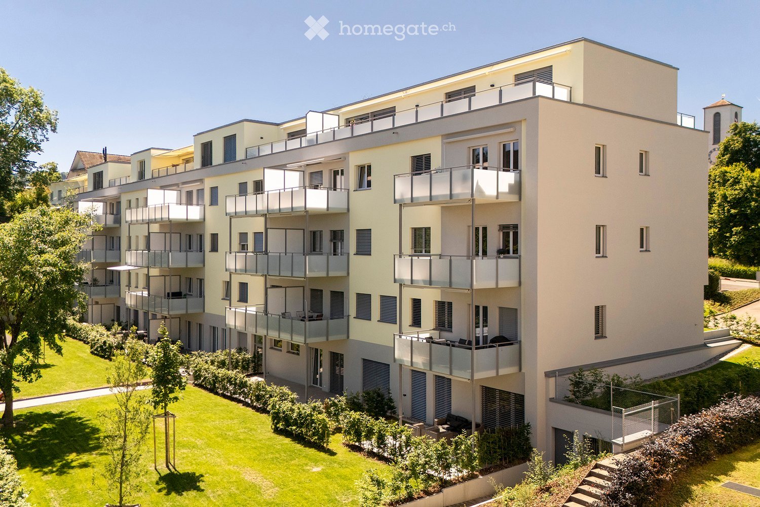Modern multi-story residential building with balconies, surrounded by green lawn and trees.