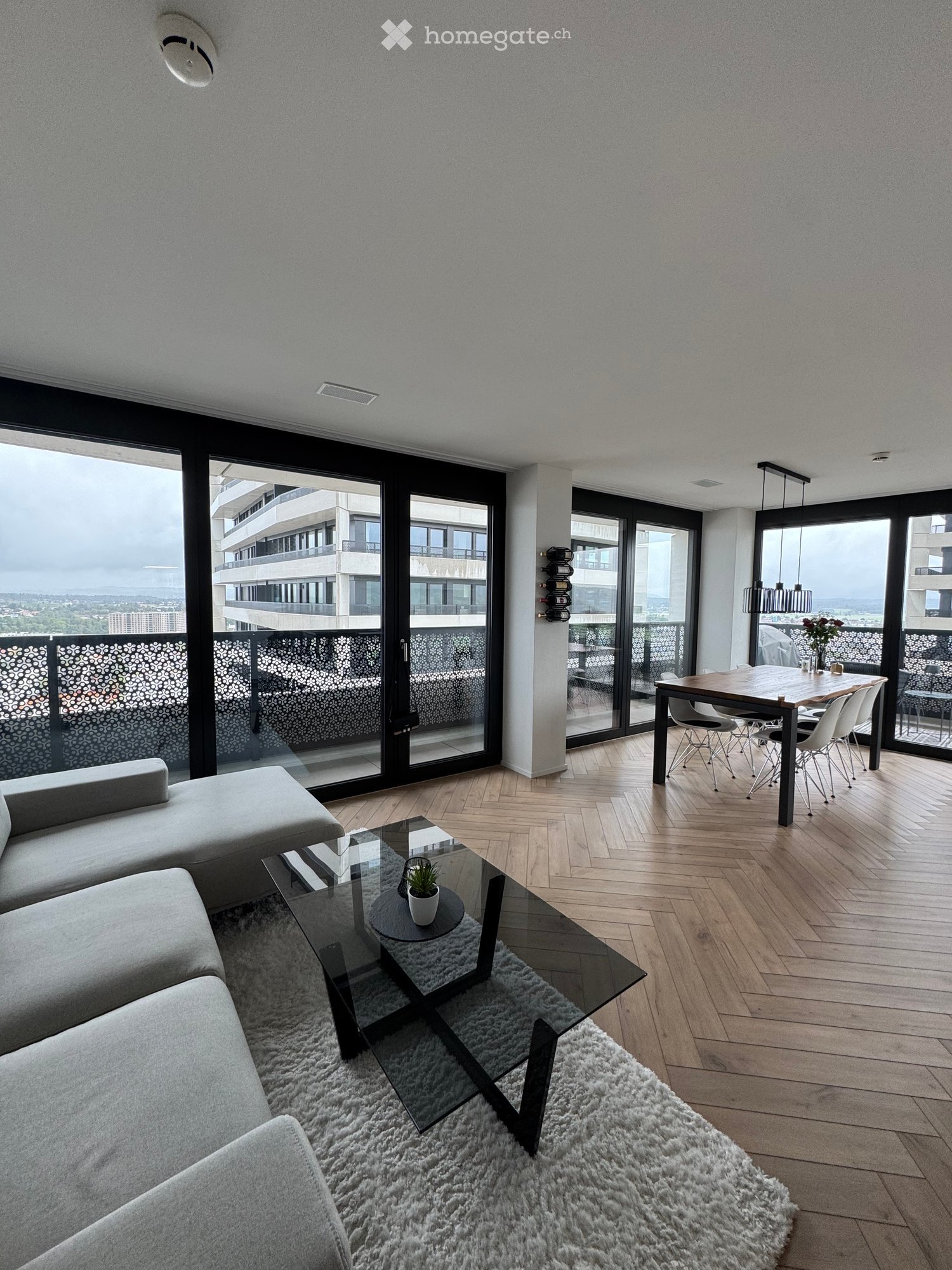 Modern living and dining area with wooden flooring, large windows offering a view, a grey sectional sofa, a glass coffee table with a small plant, a dining table with chairs, and a fluffy white rug.