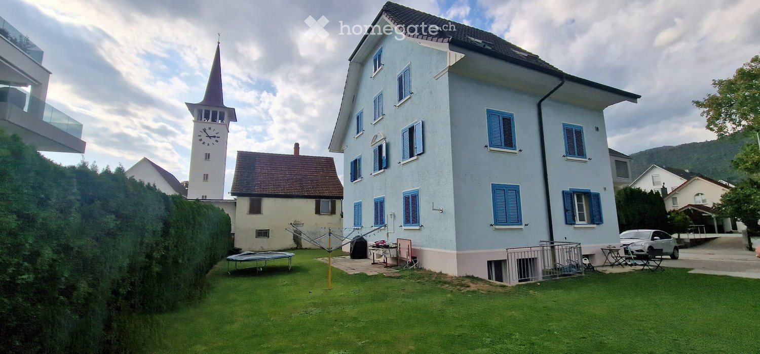 Two-story house with light blue exterior and blue shutters, surrounded by a well-maintained lawn and outdoor furniture.