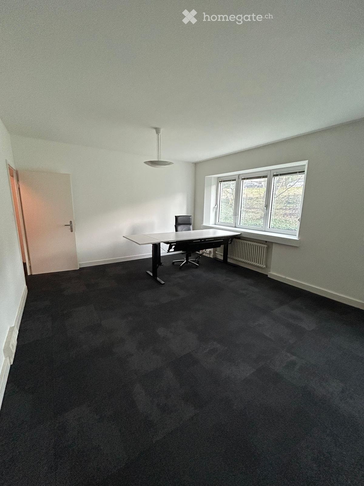 Office space with a black carpeted floor, white walls, a large window, a table, and a chair.