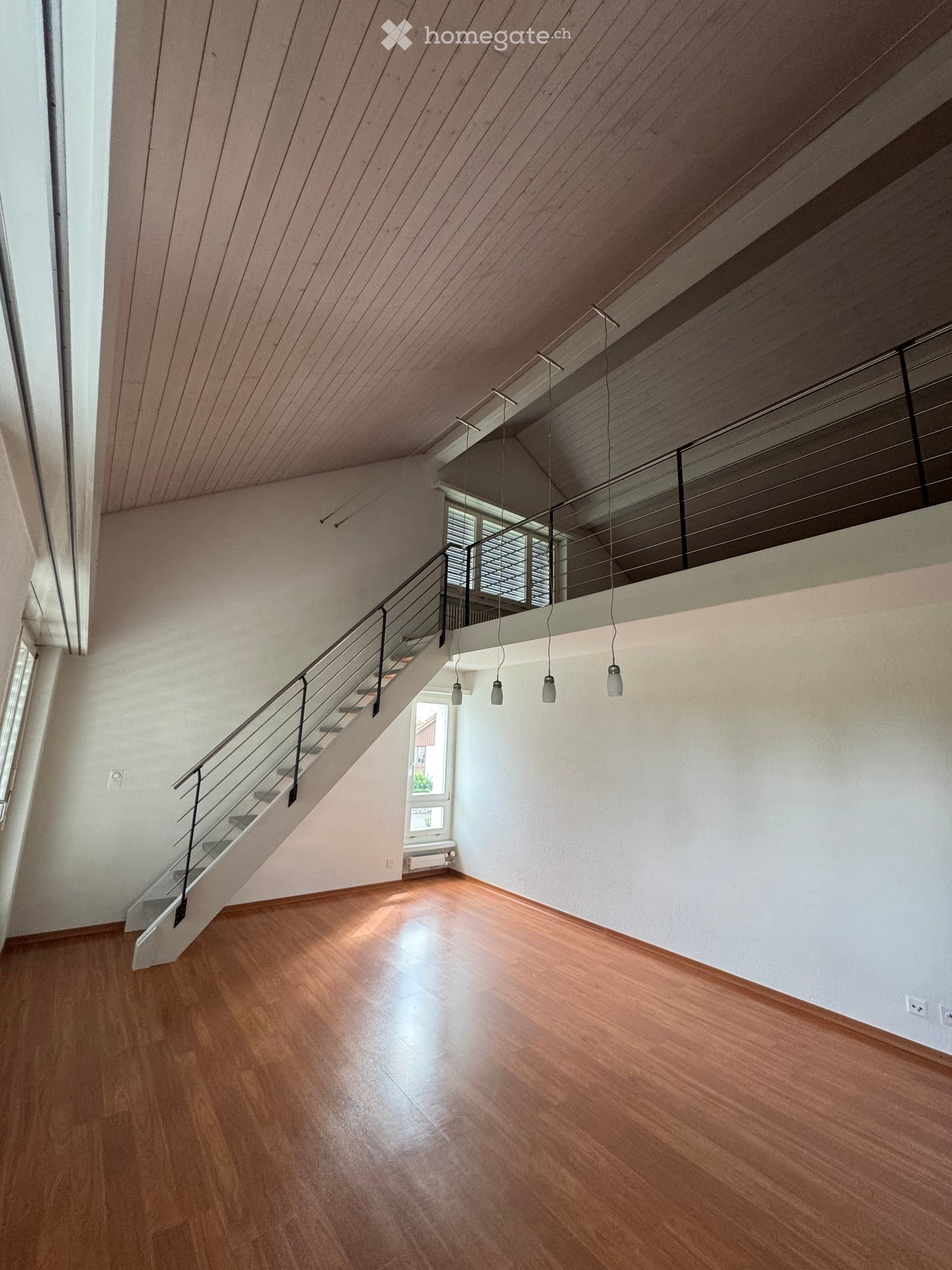 Empty room with wooden flooring, white walls, and a staircase leading to an upper level. The space features modern lighting fixtures and a sleek metal railing on the staircase.