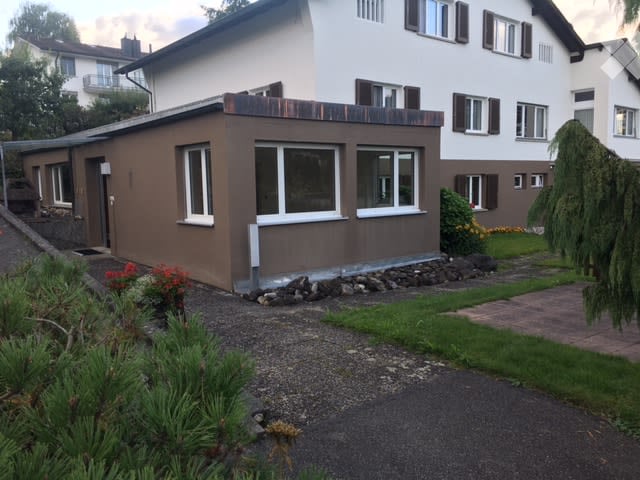 A two-story house with a modern, brown facade and white trim around the windows. The house features a flat roof, multiple windows, and is surrounded by a well-maintained garden with shrubs and a gravel driveway.