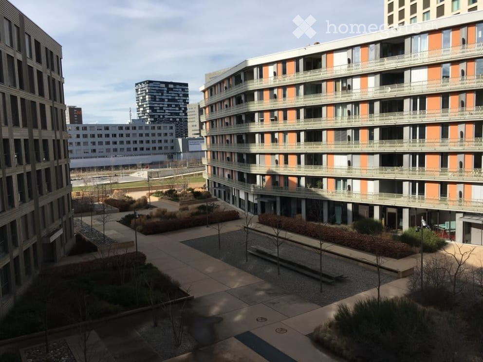 Modern multi-story residential building with balconies and contemporary design, surrounded by landscaped area with pathways and small trees.