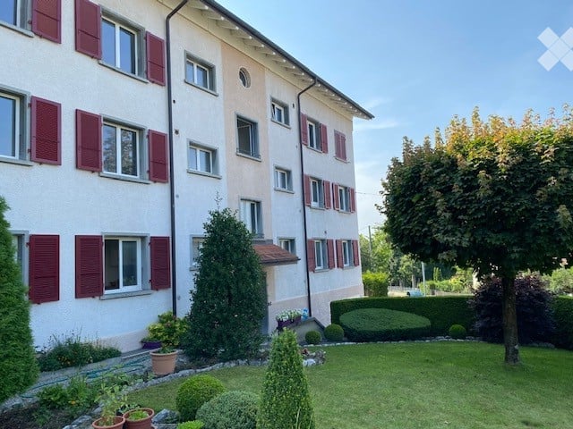Multi-story residential building with white and pink facade, red shutters, and a well-maintained garden in front.