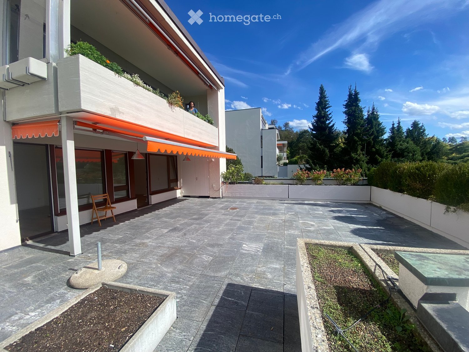 Courtyard with paved stone floor, raised planters, small bench, and modern building with balcony and orange awning.