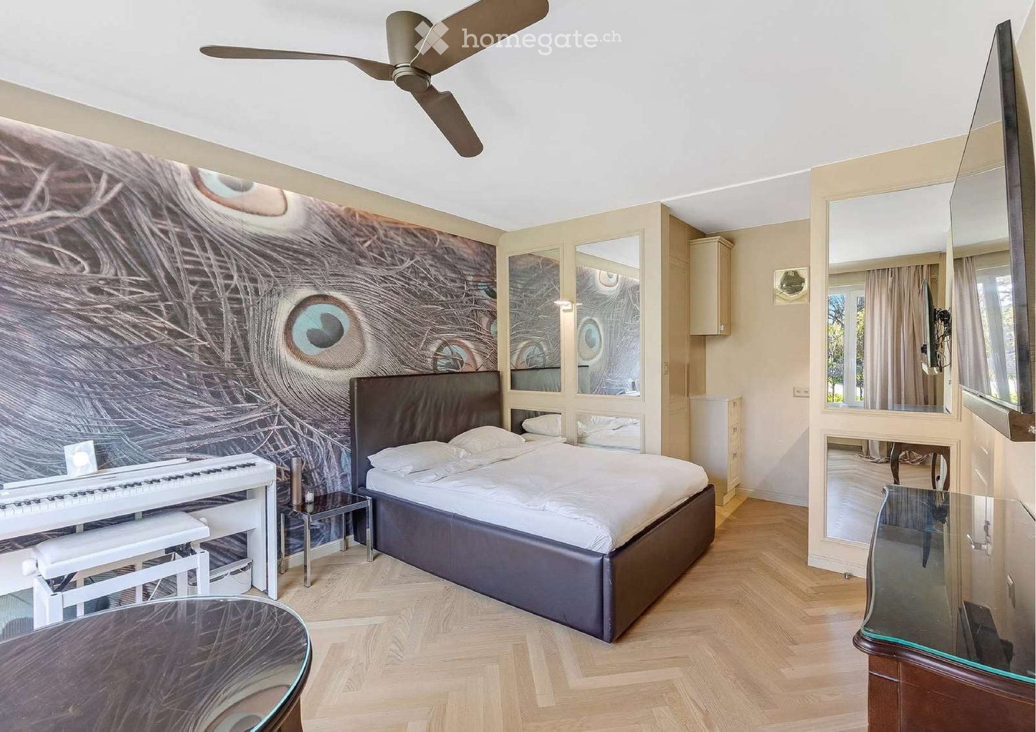 Modern bedroom with a large bed, piano, glass table, and large mirror. The walls feature an eye-themed mural.