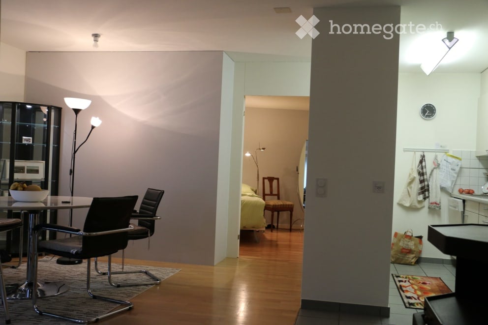 A modern, open-plan living and dining area with a sleek glass table, black chairs, a floor lamp, and a bowl of fruit. A separate bedroom is visible through an open doorway, featuring a bed, a wooden stool, and a mirror. A small kitchen area is also visibl