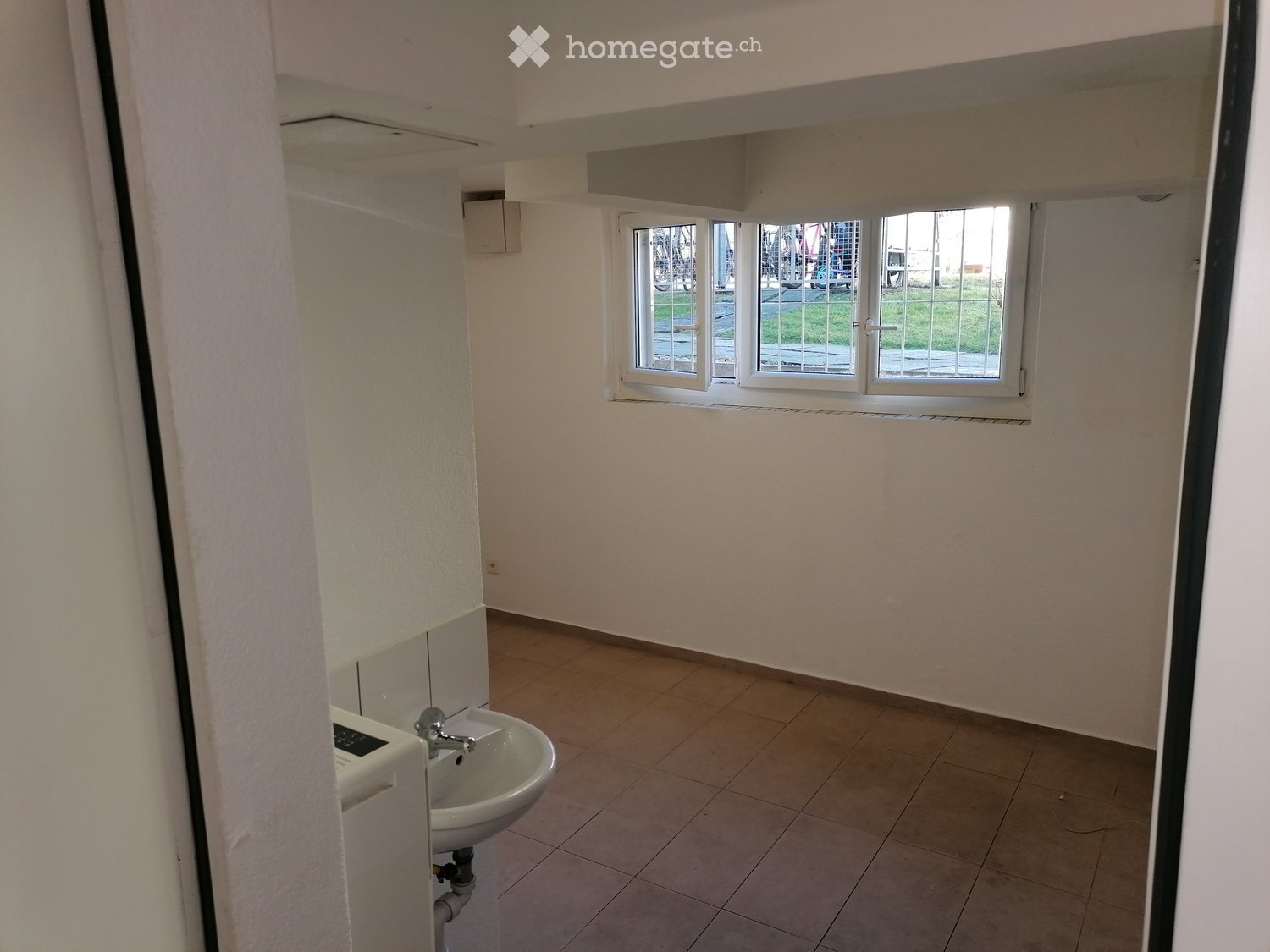 Empty bathroom with tiled floor, white walls, and a single sink. The window has bars and shows an outdoor view.