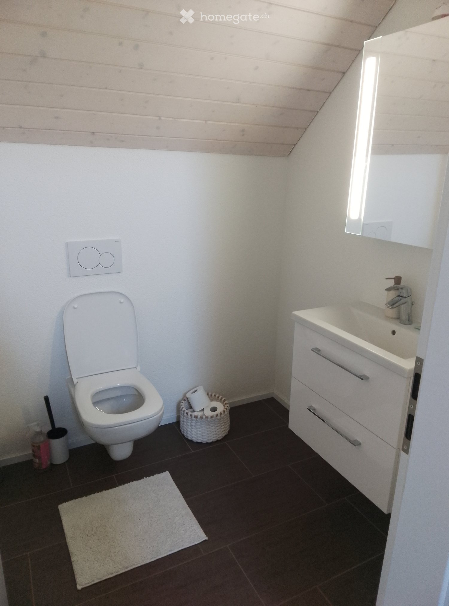 Modern bathroom with white toilet, sink, and vanity, dark tiled floor, and a small white rug.