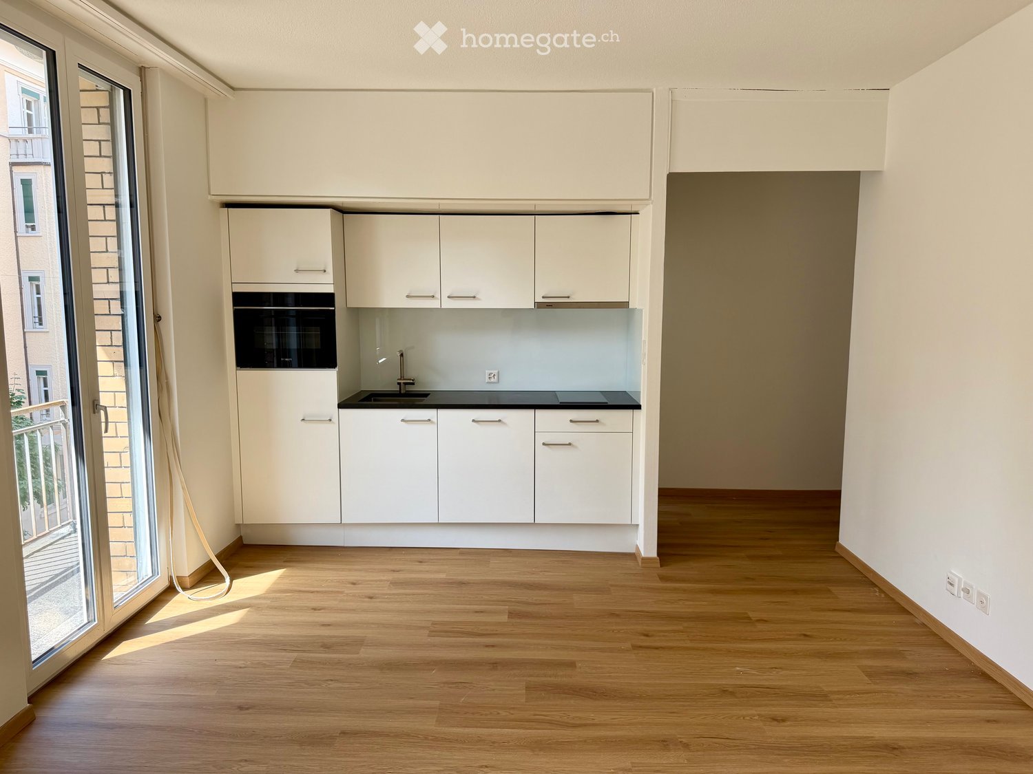 Modern kitchen with white cabinets, a sink, a faucet, and an oven, featuring wooden flooring and large windows.