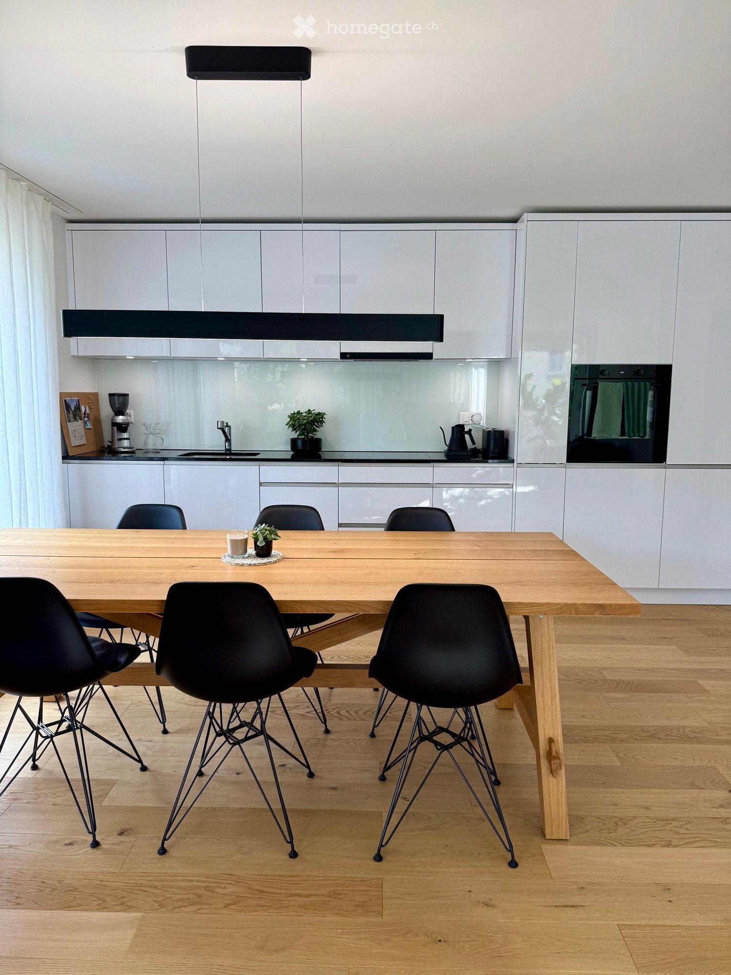 Modern kitchen and dining area with white cabinets, a wooden dining table, black chairs, and various appliances including a coffee maker, oven, and teapot.