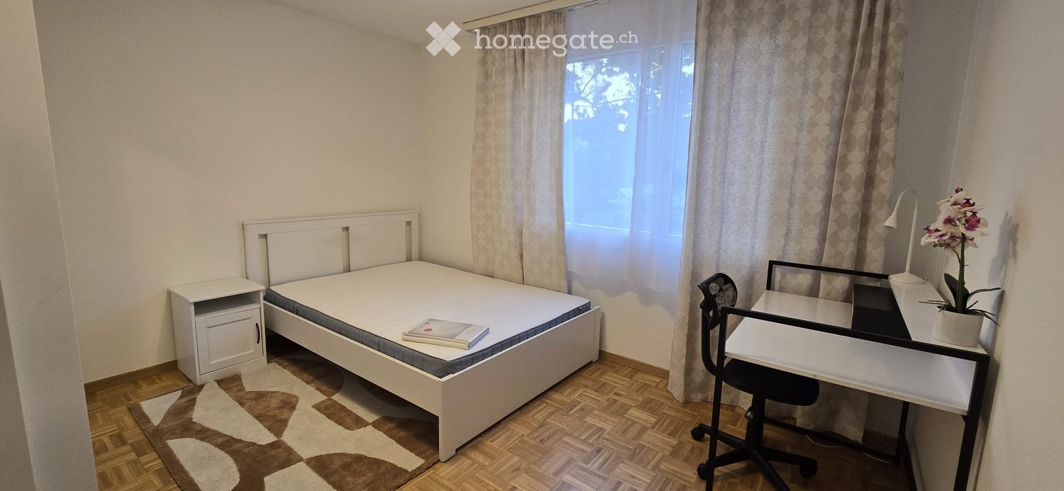Cozy bedroom with a white bed, nightstand, desk, chair, and a flower pot on the desk. The room features wooden flooring and large windows with curtains.