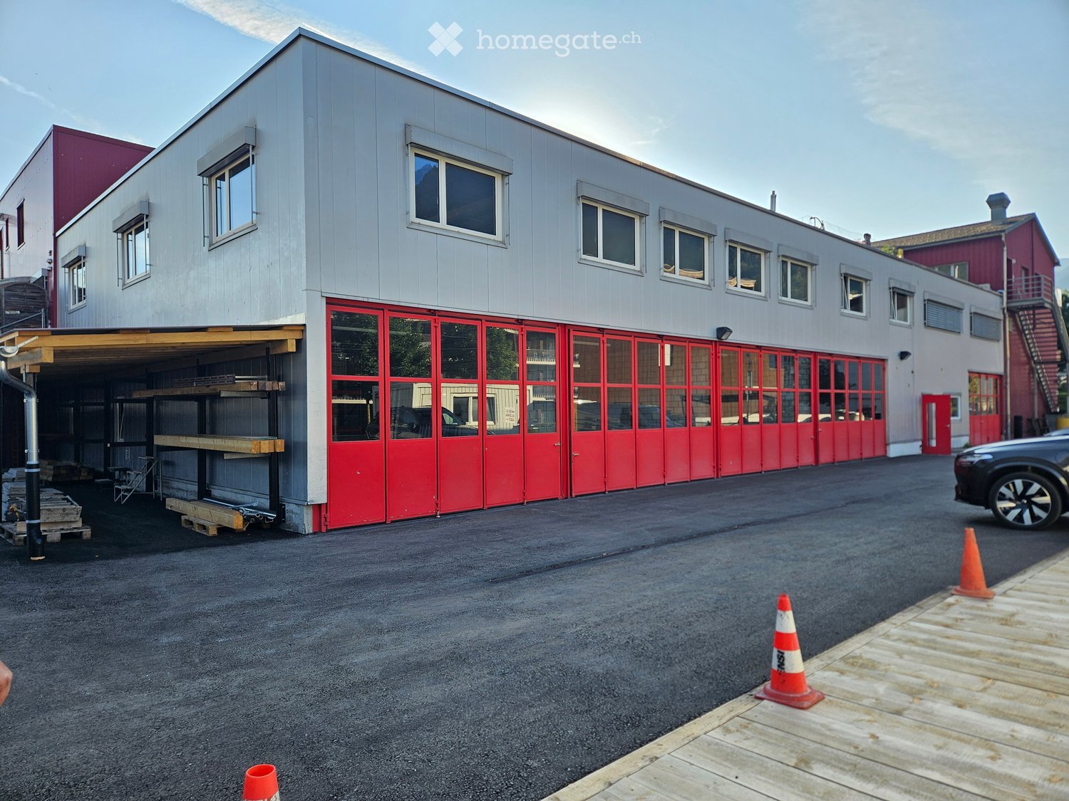 Modern two-story building with grey siding, red garage doors, and multiple windows. Includes a covered parking area and traffic cones on the asphalt ground.