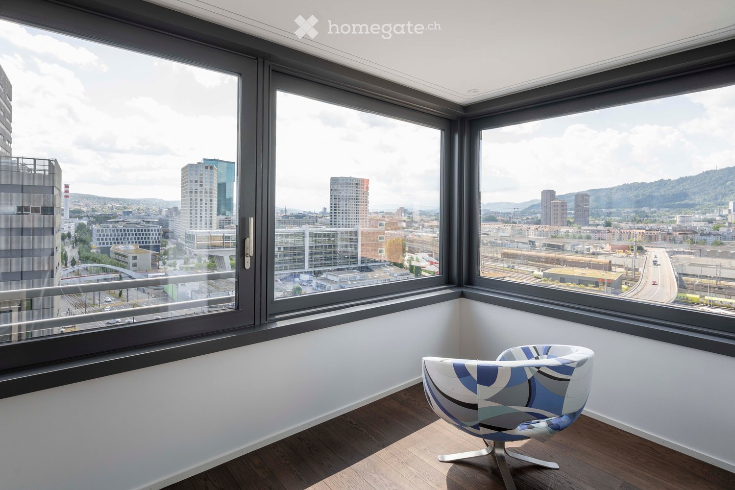 Modern room with large windows offering a cityscape view, featuring a single contemporary chair with a blue and white geometric design.