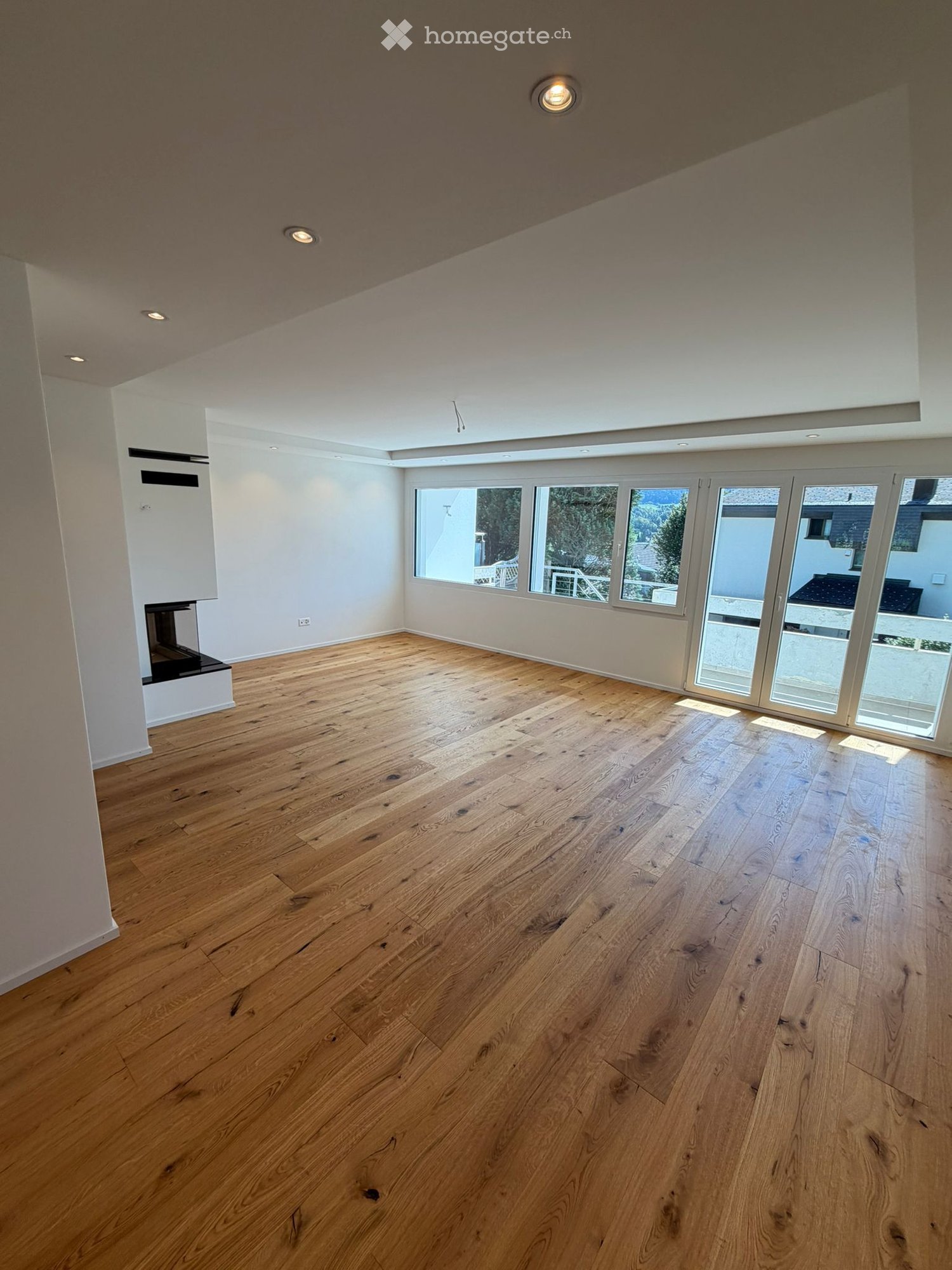 Empty room with wooden flooring, large windows, and a sliding glass door leading outside. The ceiling has recessed lighting.