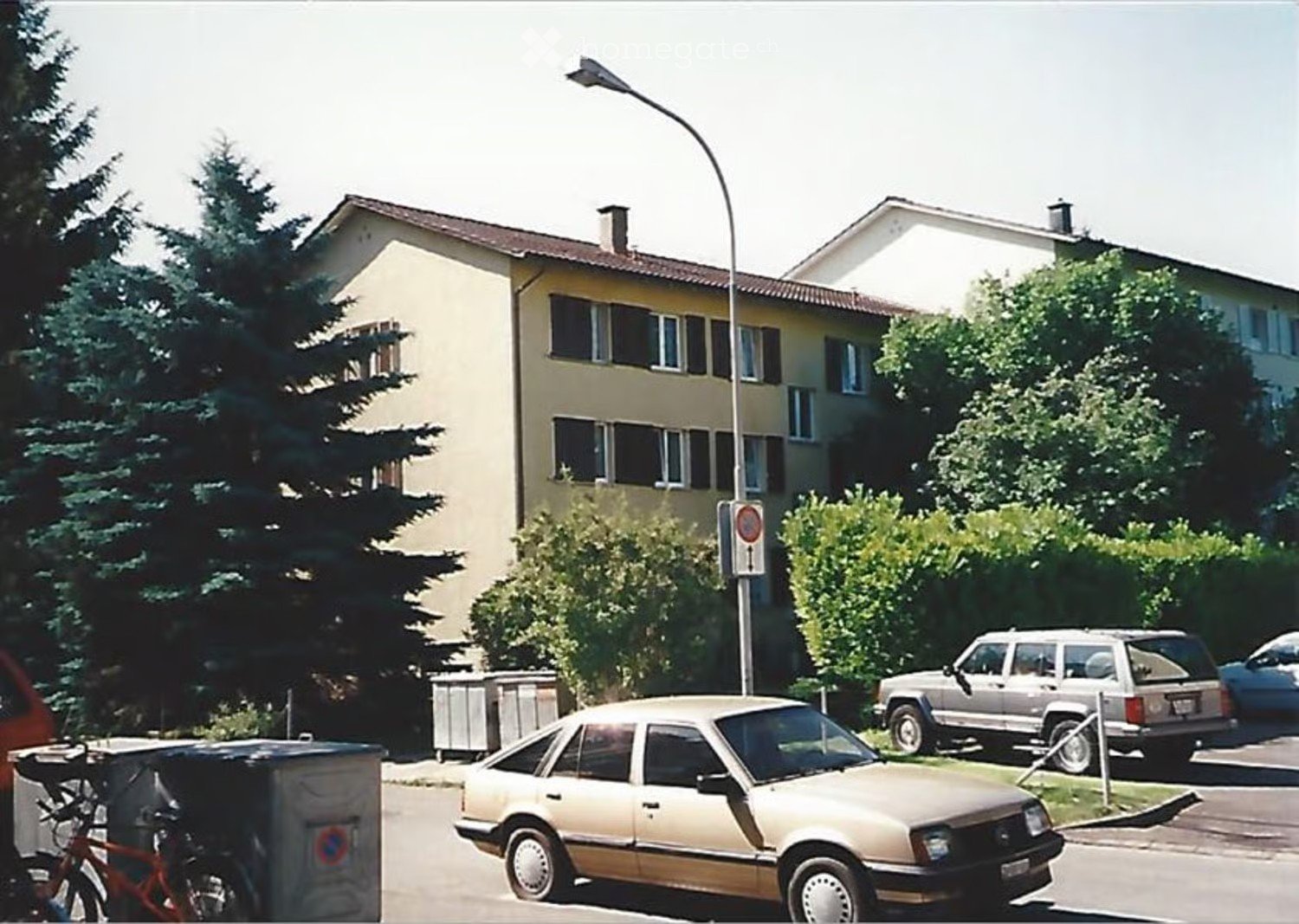 Two-story residential building with beige walls, multiple windows, and a tiled roof. The building is surrounded by trees and parked cars, with a streetlight and a stop sign visible in the foreground.