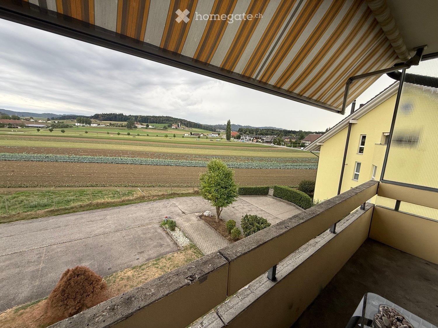 Balcony with a view of a rural landscape, including fields, a driveway, and a small garden area with shrubs and a tree. The balcony has a concrete railing and a striped awning.