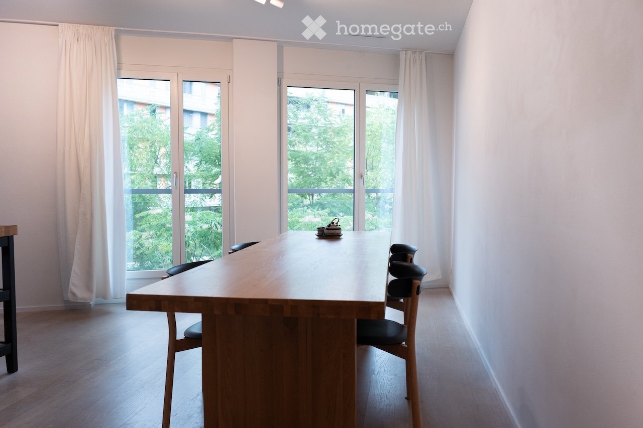 Bright dining area with a large wooden table and chairs, large windows with white curtains, and a view of greenery outside.