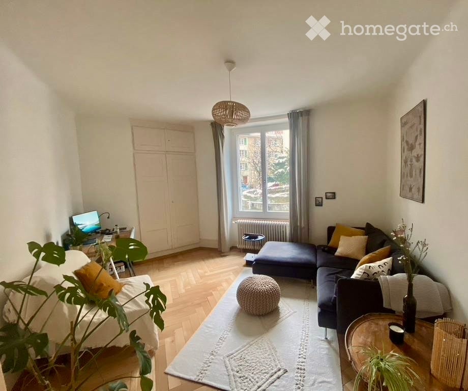 Modern living room with a sectional sofa, coffee table, ottoman, and a laptop on a desk. The room features wooden flooring, large windows with curtains, and decorative plants.