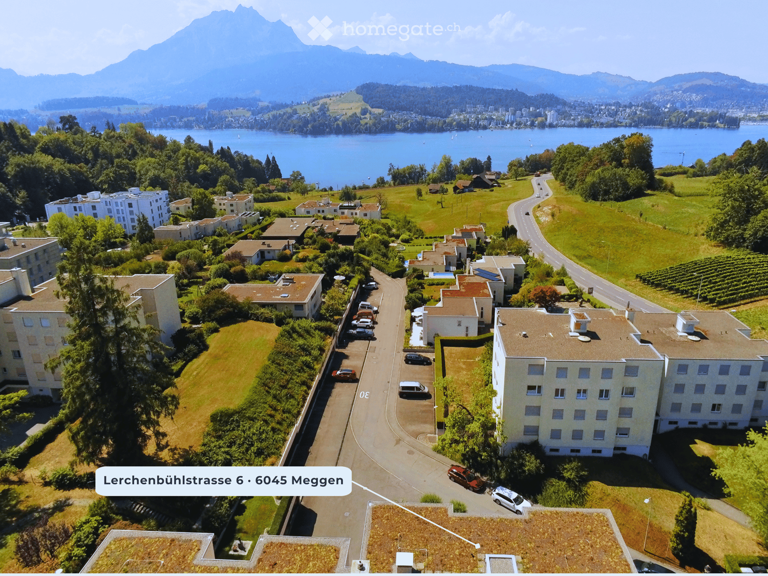 Aerial view of a residential area with multiple buildings, parking lots, and surrounding greenery, with a scenic lake and mountains in the background.