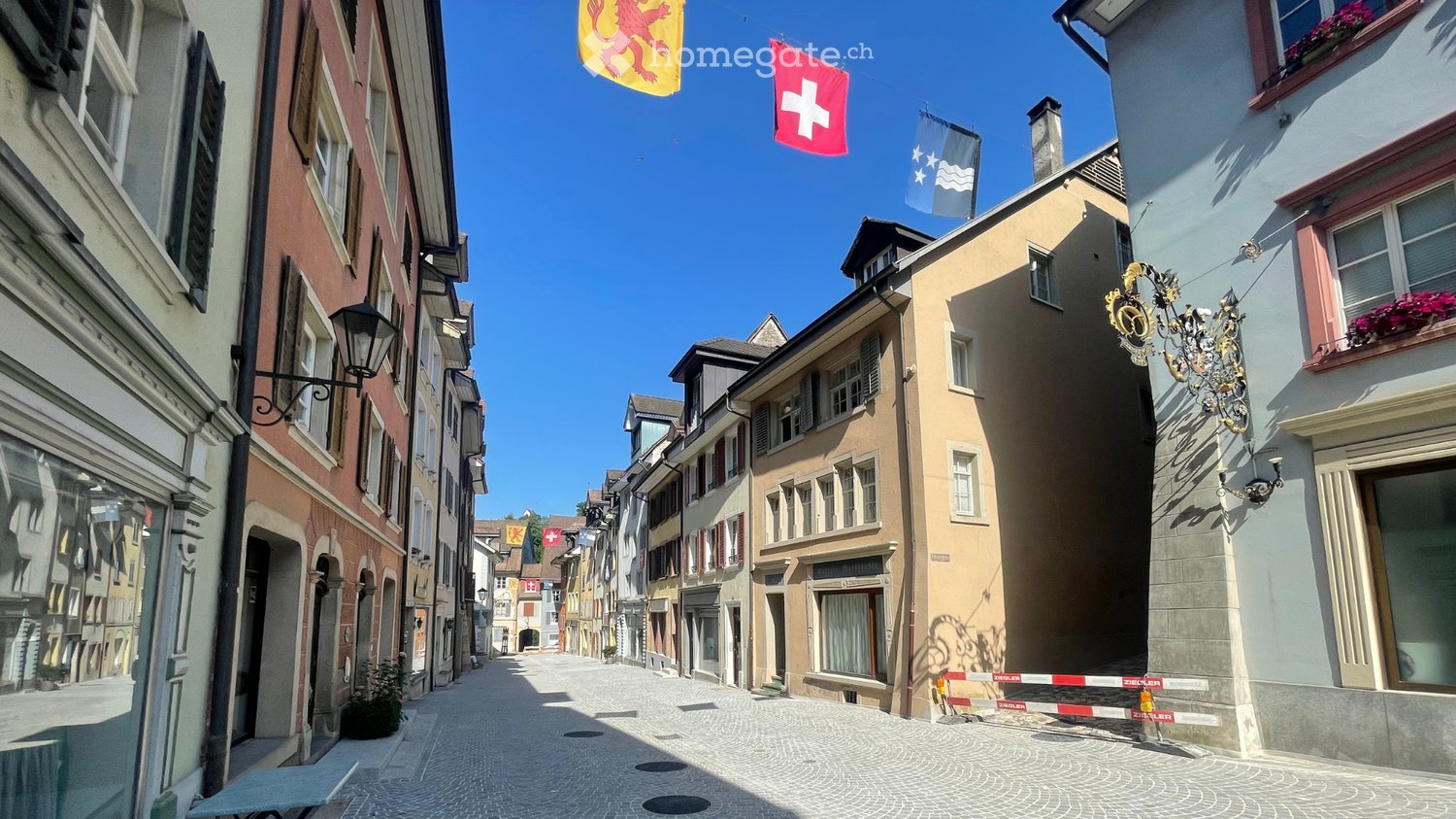 A narrow cobblestone street lined with colorful buildings, featuring traditional European architecture, flags, and ornate street lamps.