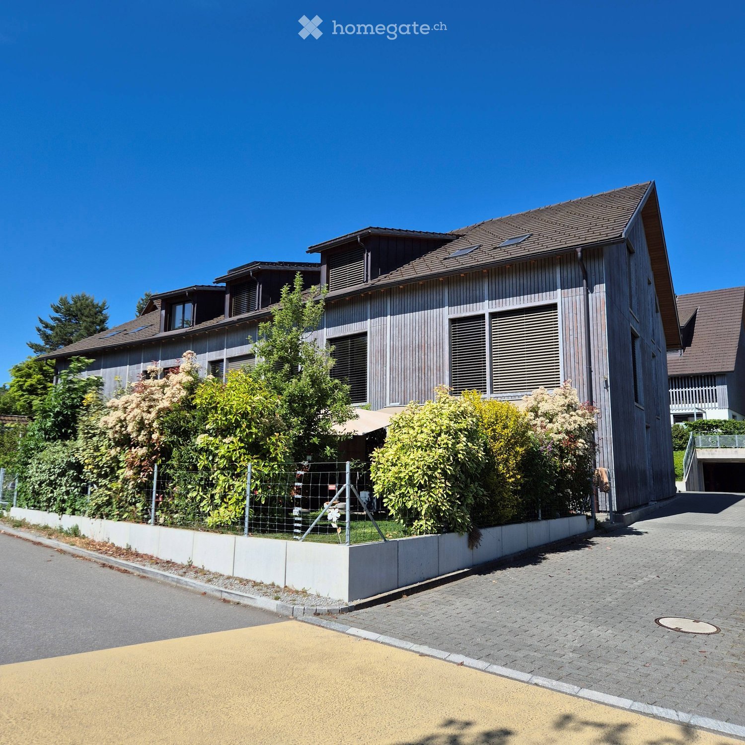 Modern two-story house with wooden exterior, multiple windows, and a fenced garden area.