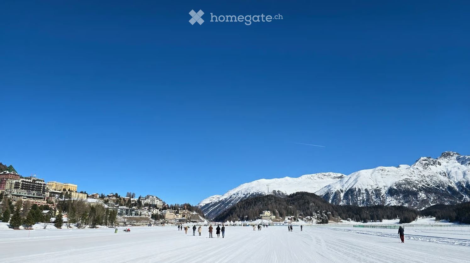 A snowy outdoor area with people walking, surrounded by snow-capped mountains and buildings in the background.