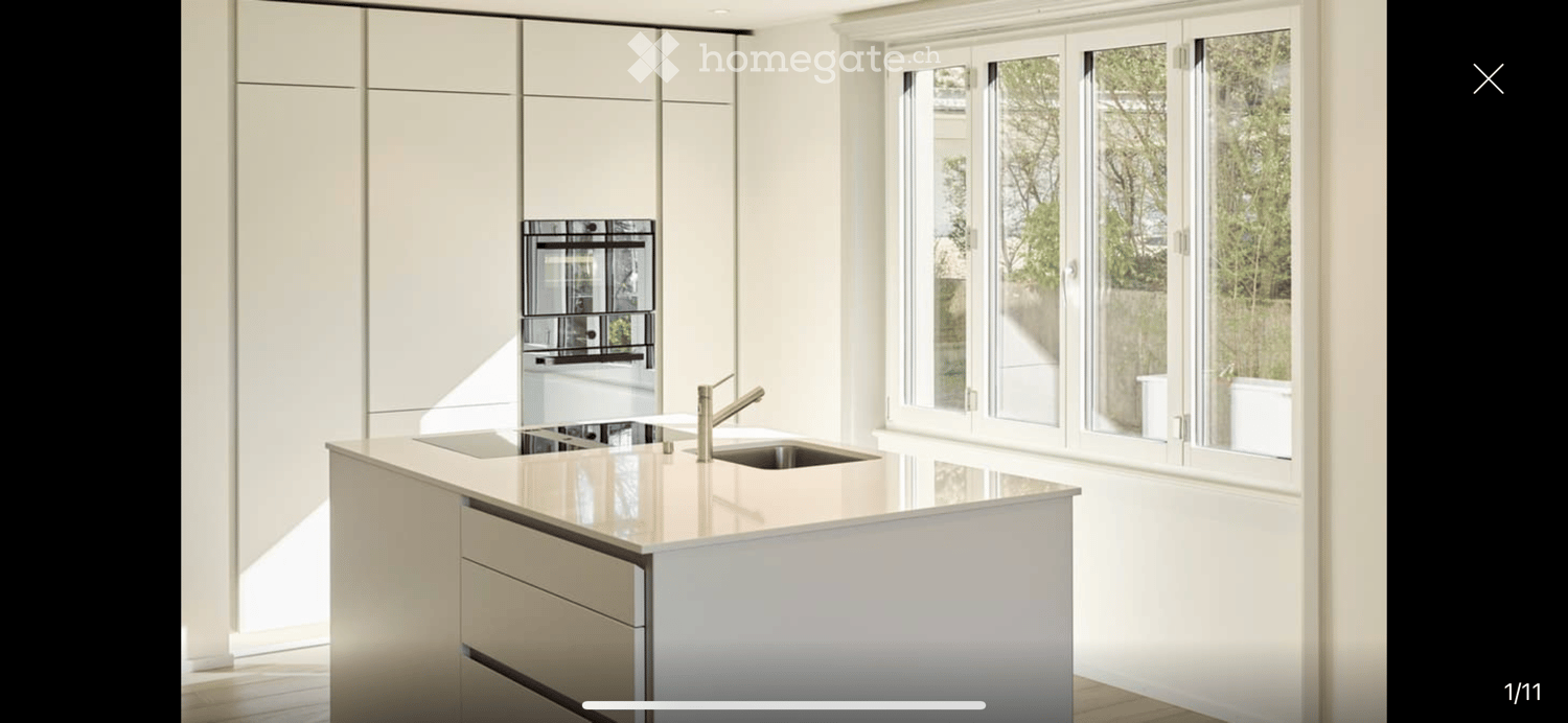 Modern kitchen with white cabinets, a central island, a double sink, and an integrated oven. Large windows provide natural light.