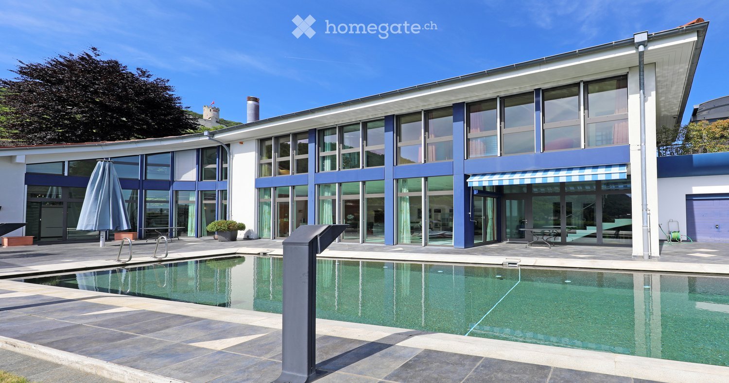 Modern two-story building with large glass windows and blue accents, featuring a swimming pool in the foreground and outdoor seating area.