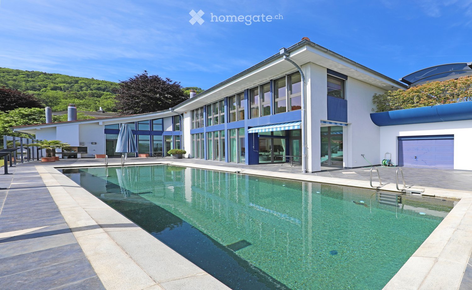Modern two-story house with large windows, a swimming pool, and a garage. The house features a sleek design with white and blue accents, surrounded by greenery and a clear blue sky.
