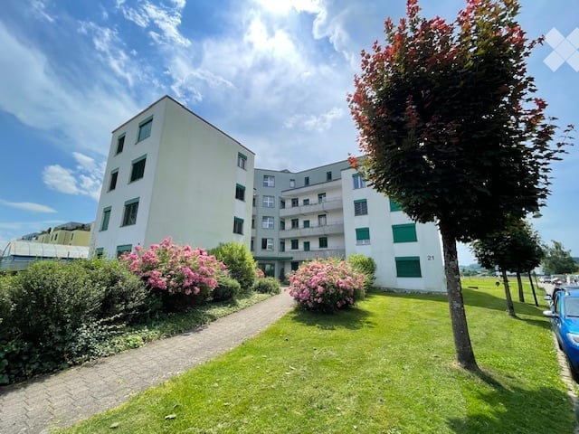 Modern multi-story residential building with white facade, green shutters, and surrounding garden area with flowering bushes and trees.