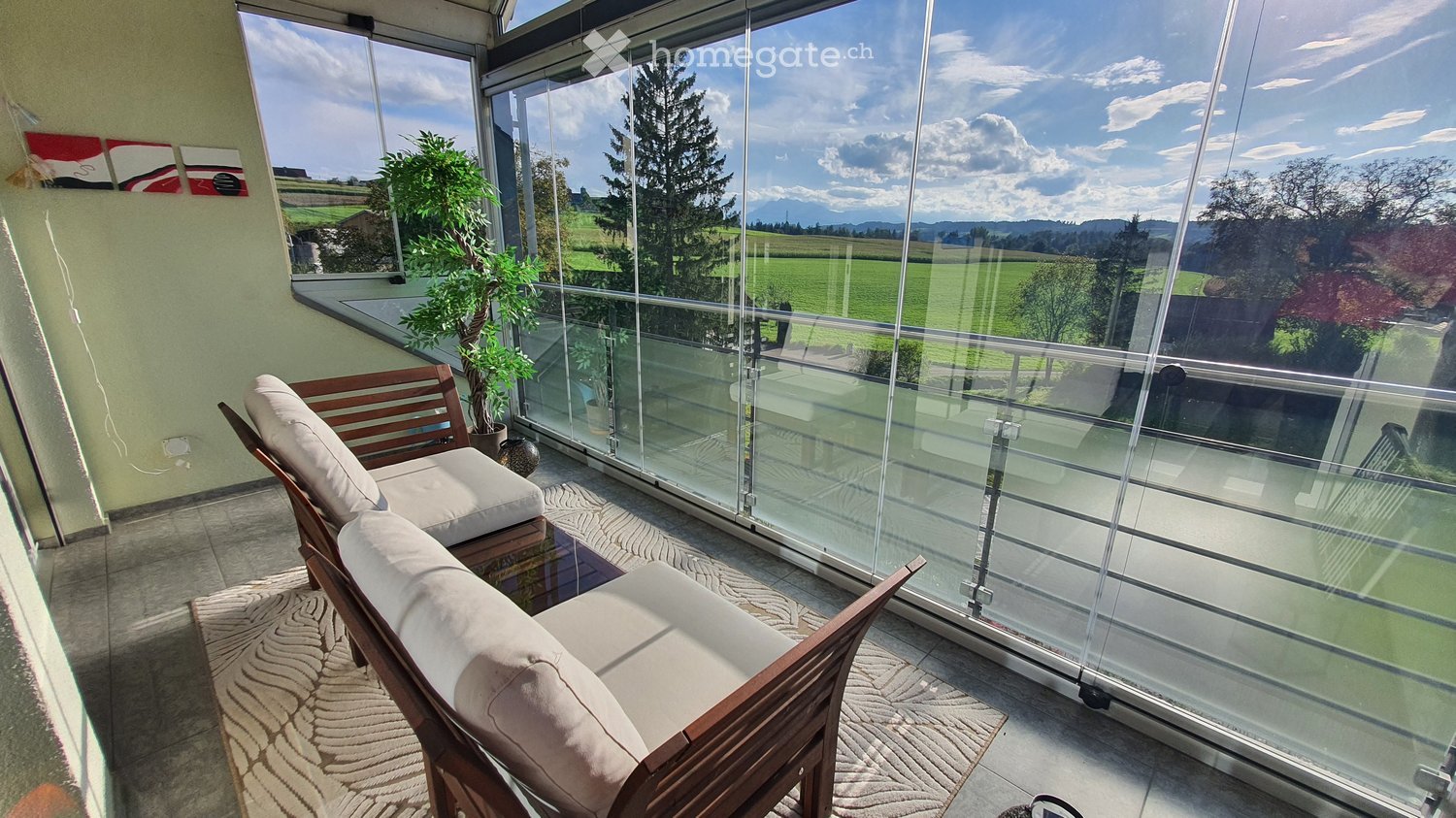 Balcony with glass railing, wooden sofa, small table, and potted plant, overlooking a scenic countryside view.