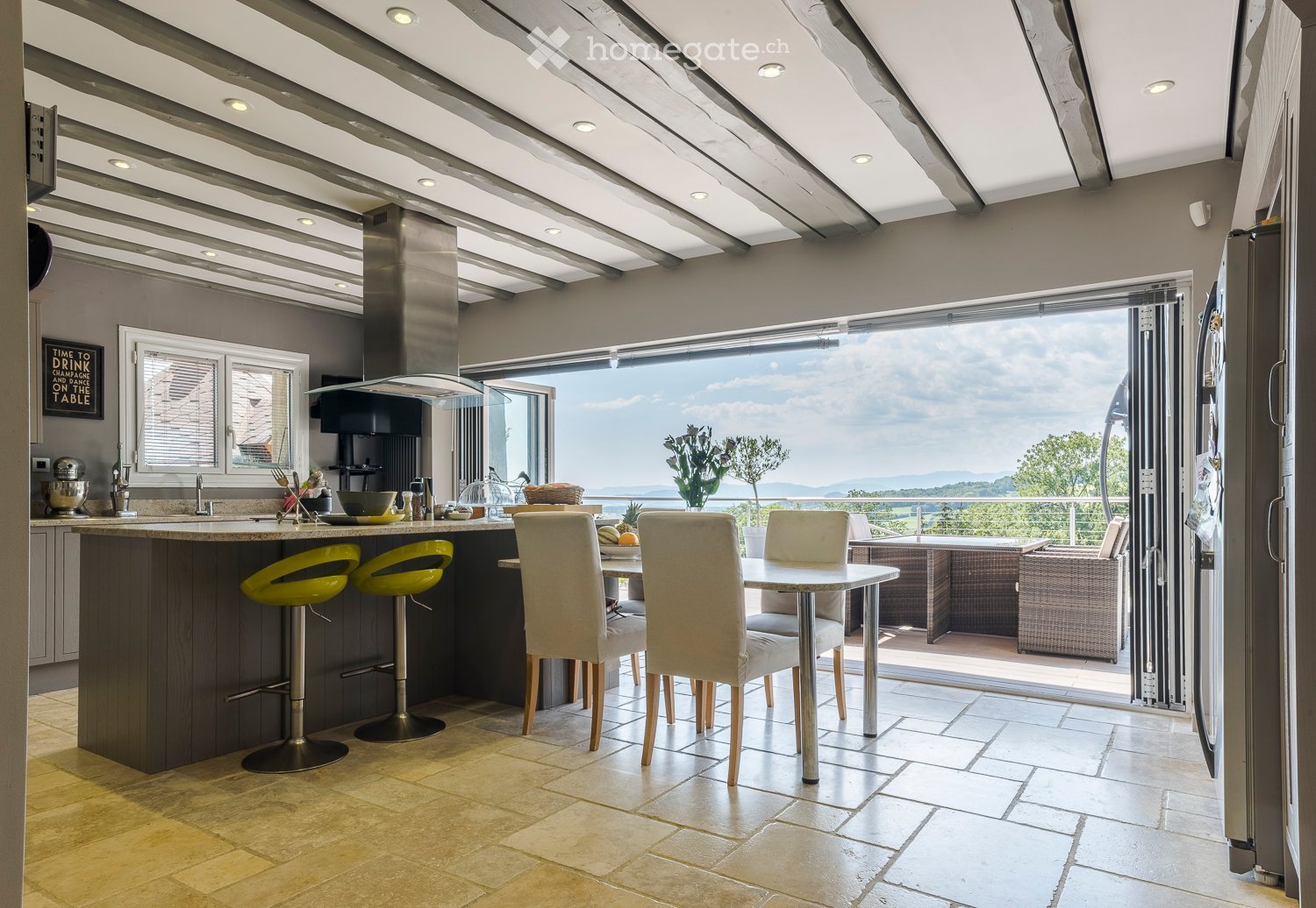 Modern kitchen with island, bar stools, dining table, chairs, and a view of the landscape through large windows.