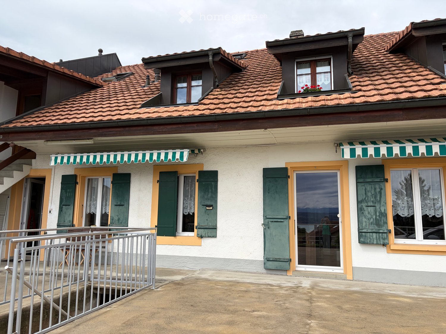 Two-story house with red-tiled roof, white walls, green shutters, and striped awnings over the windows.