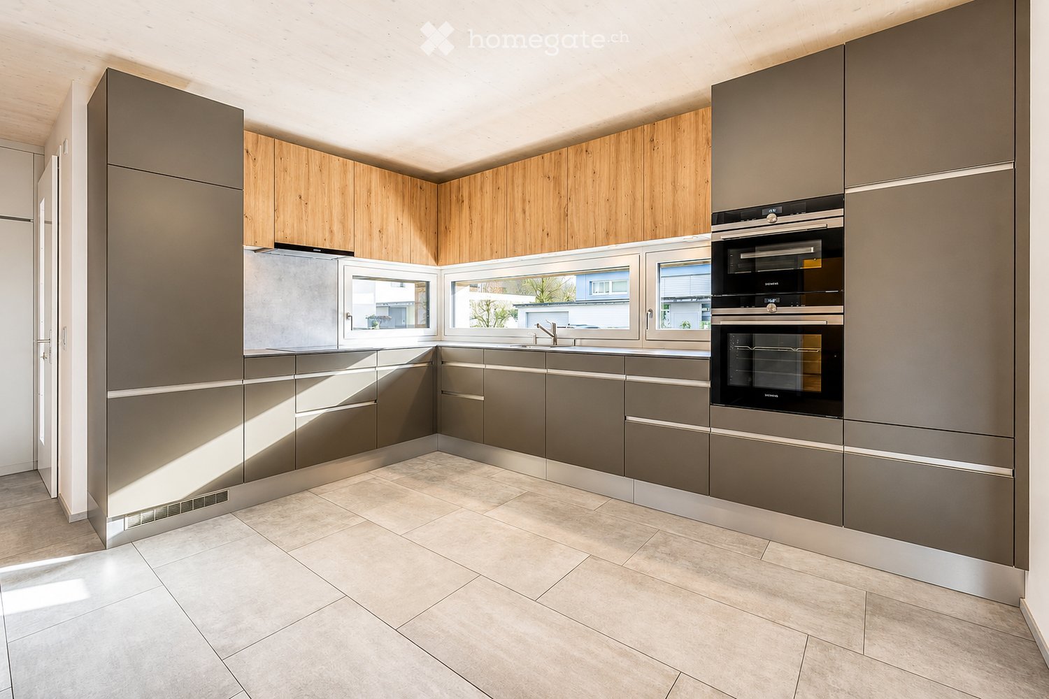 Modern kitchen with grey and wooden cabinets, stainless steel appliances, and large windows letting in natural light.