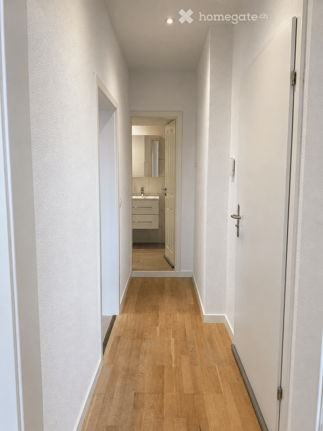 A narrow hallway with light wooden flooring and white walls, leading to a small bathroom with a sink and mirror.