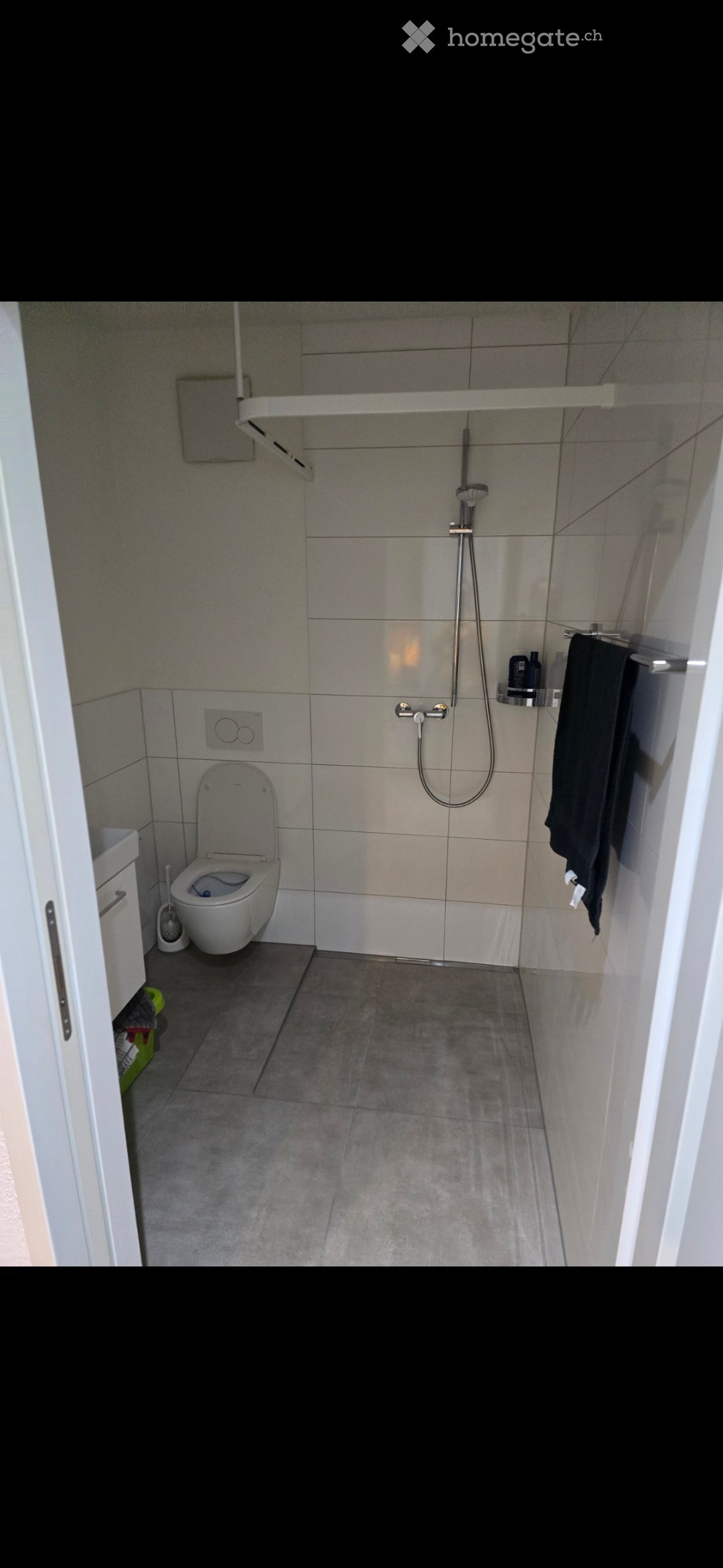 Modern bathroom with a toilet, shower, towel, and shower shelf. The walls are tiled in a light gray color, and the floor is covered with dark gray tiles.