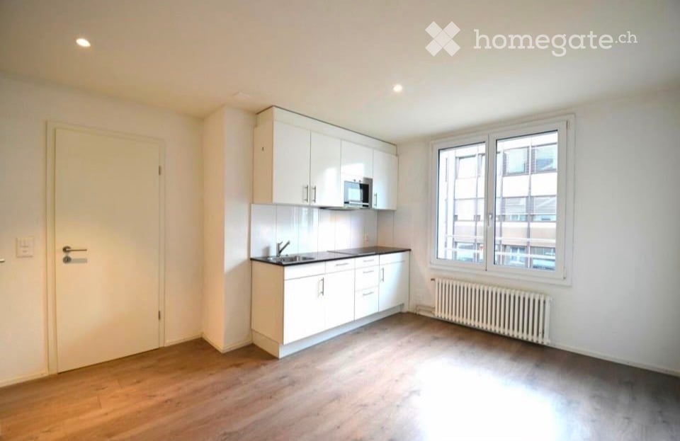 Empty kitchen with white cabinets, a microwave, sink, and countertop. Wooden flooring and large windows.
