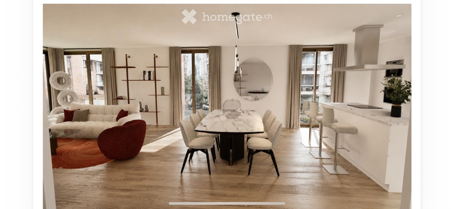 Open-plan living and dining area with white walls, wooden flooring, large windows, a white sofa, a round red chair, a marble dining table with chairs, a kitchen island with bar stools, and decorative shelves.