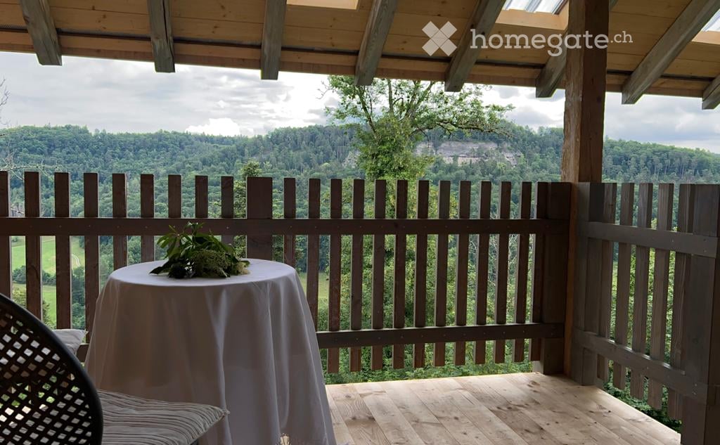 Wooden balcony with a scenic mountain view, featuring a small round table with a floral centerpiece and a wicker chair.
