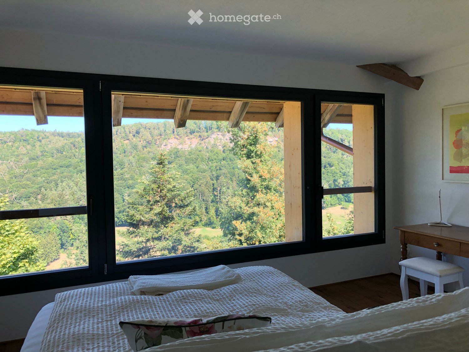 Bright bedroom with large windows offering a scenic view of a forested landscape, featuring a bed with patterned bedding, a wooden desk, and a white stool.