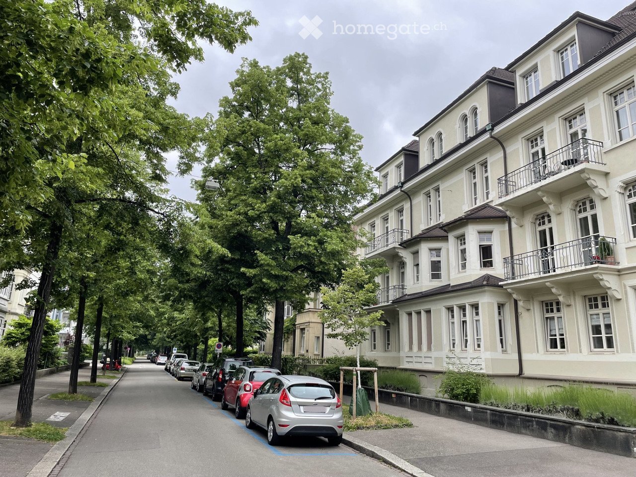 A multi-story residential building with balconies and large windows, situated on a tree-lined street with parked cars.