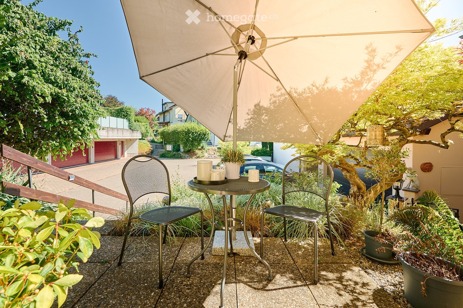 Balcony with a small round table, two metal chairs, and a large umbrella providing shade. Potted plants and a view of the street are visible.