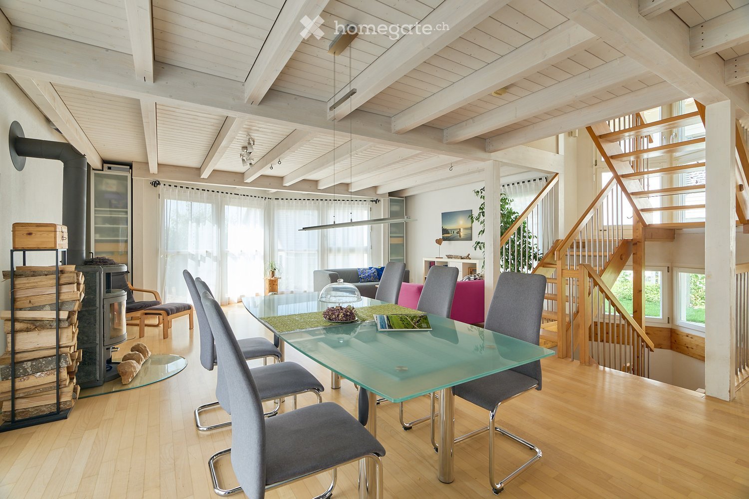 Bright, open-plan living and dining area with wooden floors, exposed wooden beams, a glass dining table with six chairs, a sofa, an armchair, a fireplace with a log rack, a wooden staircase leading to an upper level, and large windows with white curtains.