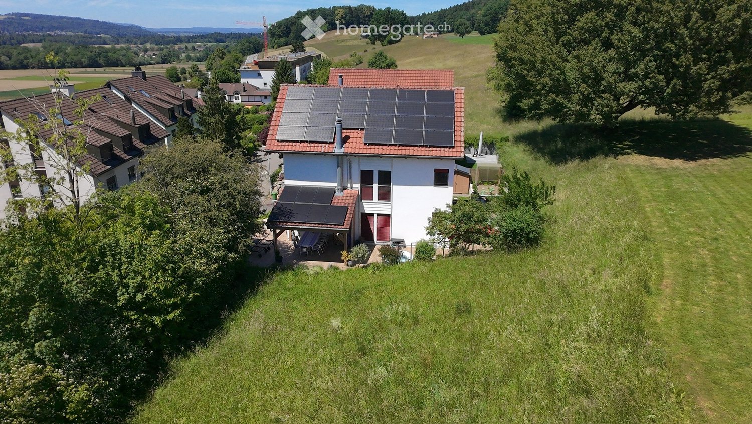 A modern two-story house with solar panels on the roof, surrounded by a well-maintained lawn and trees. The house features white walls, red shutters, and a covered patio area with outdoor furniture.